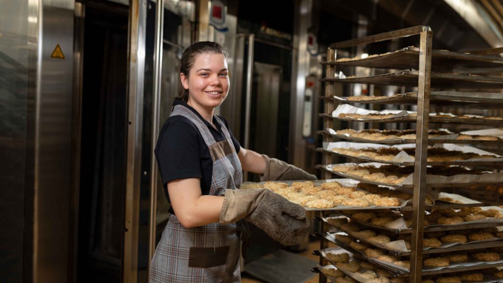 Junges Mädchen beim Projekt "Der soziale Tag" in einer Bäckerei. Sie schiebt gerade ein Blech mit Keksen auf den Wagen.