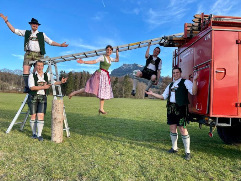 Elbach feiert den Brass Freida und Bavarian Ninja im Festzelt Elbach. Gruppe von jungen Trachtler aus Elbach stehen vor altem Feuerwehrauto und schwingen sich an einer Leiter entlang. Sie symbolisieren das Event "Bavarian Ninja".